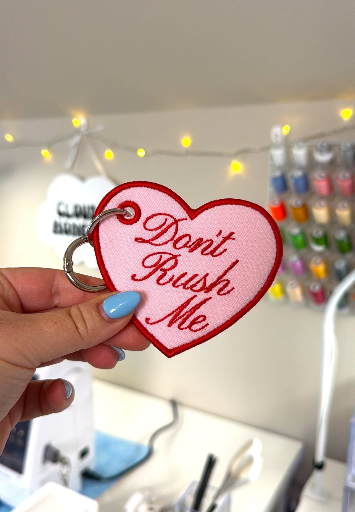 A hand with blue nail polish holds a pink heart-shaped keychain with red lettering that says 'Don't Rush Me' against a blurred desk background with string lights.