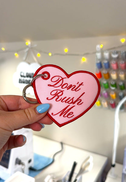 A hand with blue nail polish holds a pink heart-shaped keychain with red lettering that says 'Don't Rush Me' against a blurred desk background with string lights.