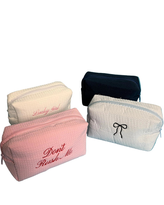 Four textured zippered cosmetic pouches arranged on a blue background: one white with pink script reading 'Lucky Girl,' one pink with red script reading 'Don't Rush Me,' one light blue with a navy blue bow motif, and one dark navy.