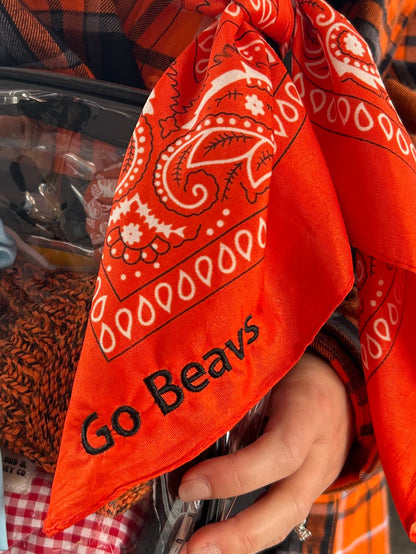 A close-up of a person holding a bright orange bandana with a white paisley pattern and the text 'Go Beavs' printed in black. The person is wearing an orange and black plaid shirt, and other items are visible in the background.