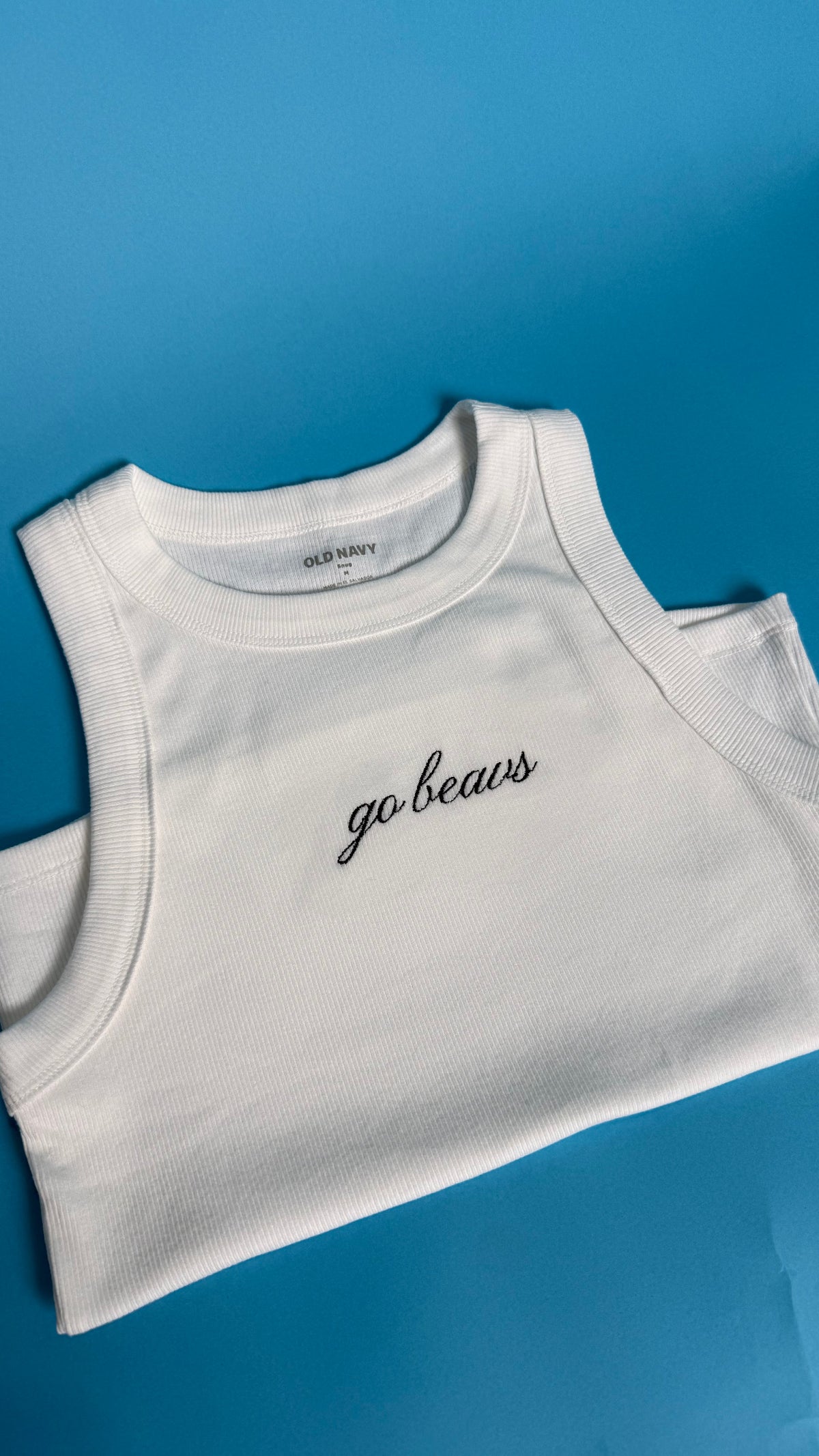A white ribbed tank top with 'go beavs' embroidered in black script on the chest, laid flat on a bright blue background.