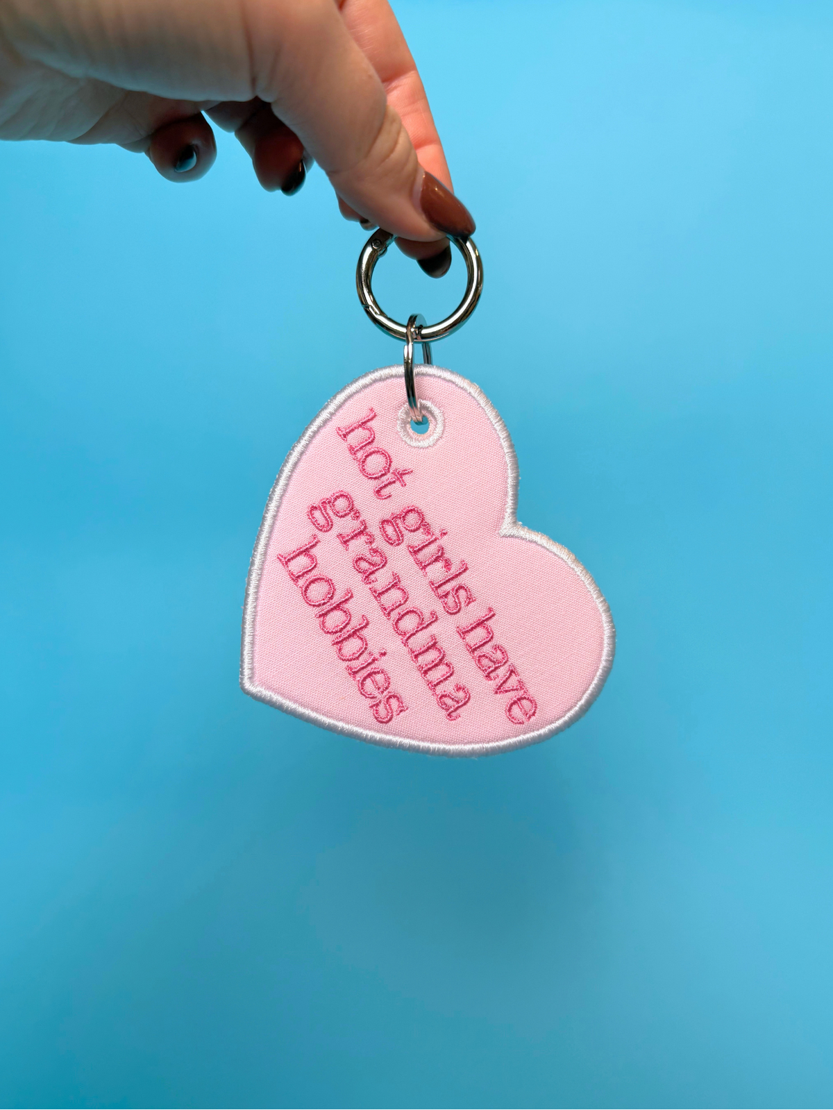 A hand holding a light pink heart-shaped keychain with embroidered text that reads 'hot girls have grandma hobbies' in darker pink. The keychain has a silver metal ring and is set against a solid bright blue background.