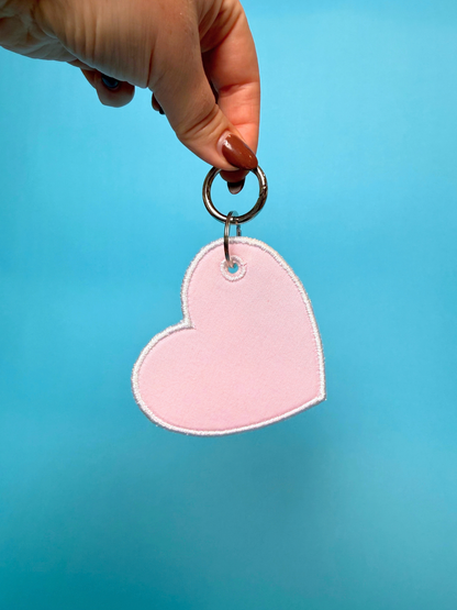 A hand with brown nail polish holds a light pink felt heart-shaped keychain with a silver metal key ring against a solid blue background.