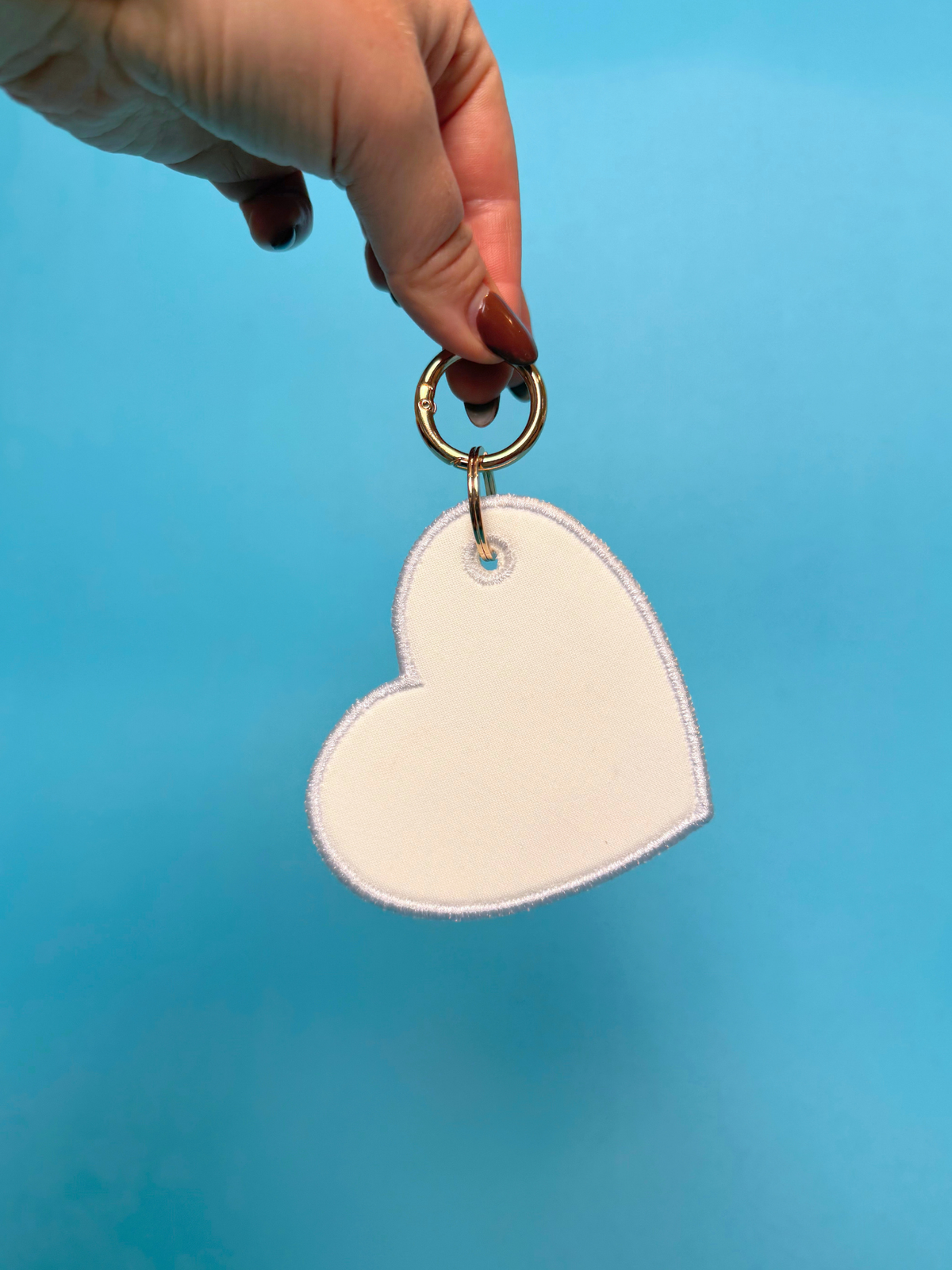 A hand with dark nail polish holds a white heart-shaped keychain with a gold metal ring against a solid blue background.