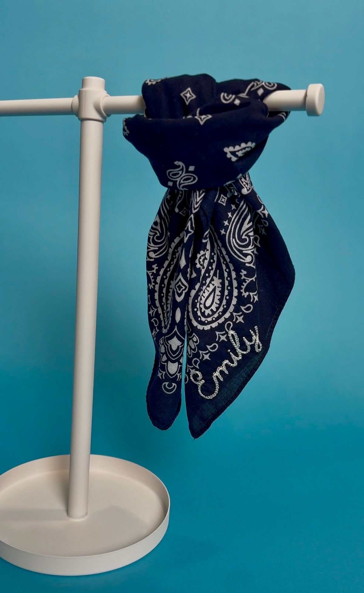 A navy blue bandana with a white paisley pattern and the name 'Emily' embroidered in white script, hanging from a white plastic display stand against a solid blue background.