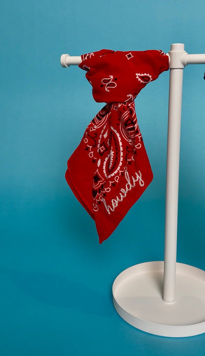 A red bandana with white paisley patterns and the word 'howdy' in script, draped over a white display stand against a solid blue background.
